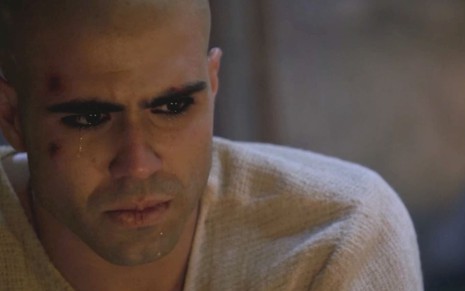 Juliano Laham em cena de Gênesis: ator está com o rosto machucado, cabelo raspado e túnica marrom clara
