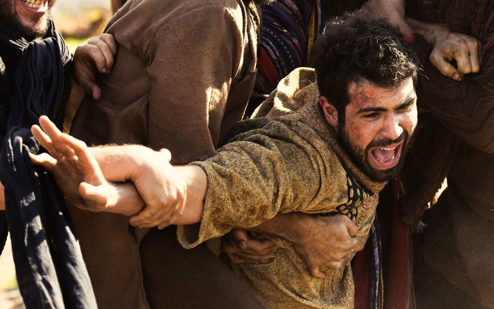 Juliano Laham em cena de Gênesis: ator está caracterizado com túnica marrom, grita e é segurado por homens que estão fora do quadro