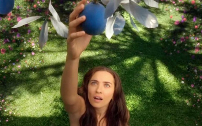 A atriz Juliana Boller como Eva está nua, com a câmera focando a partir de seus ombros, e estende o braço para pegar uma fruta redonda e azul em uma árvore em cena de Gênesis