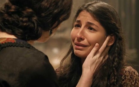 A atriz Juliana Xavier com expressão de choro e lágrimas nos olhos como Tamar, a mão de uma mulher em seu rosto a consolando, em cena de Gênesis