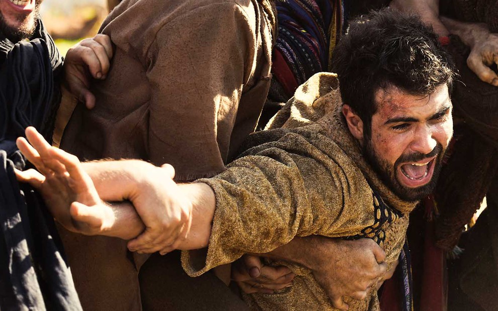 O ator Juliano Laham com o rosto machucado é agarrado por três homens que fazem menção de jogá-lo em um poço em cena de Gênesis