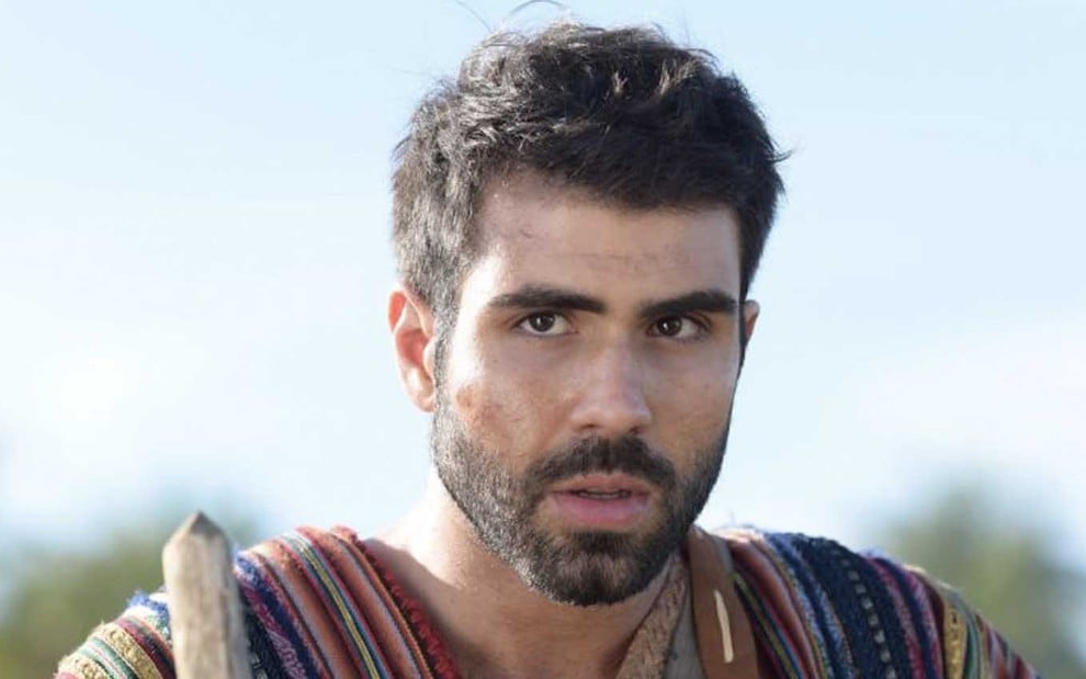 O ator Juliano Laham está com uma túnica de várias cores, com o cabelo curto e barba rente ao rosto, enquanto segura um cajado em cena de Gênesis