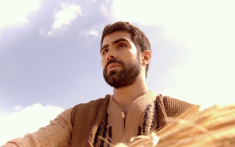 O ator Juliano Laham carrega um feixe de trigo com o céu nublado ao fundo como José em uma das cenas de Gênesis