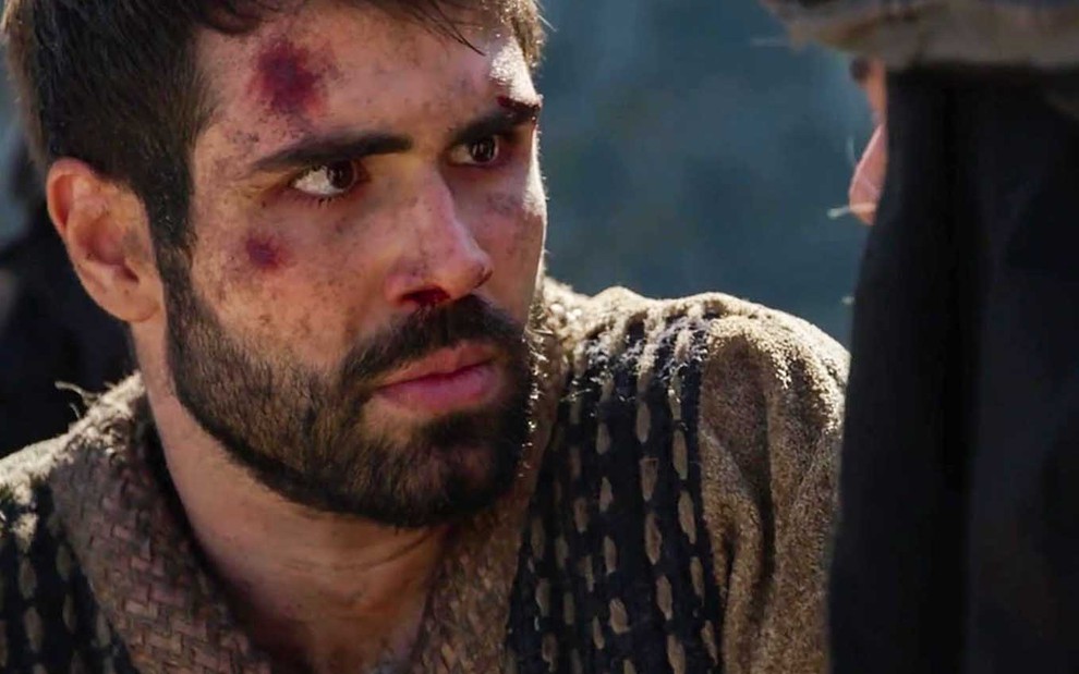 O ator Juliano Laham como José com o rosto com vários machucados olha com espanto para um homem de costas à direita em cena de Gênesis