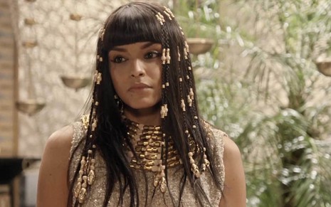 A atriz Kizi Vaz com uma peruca de cabelos pretos adornada com várias peças de ouro como a Kamesha em cena de Gênesis