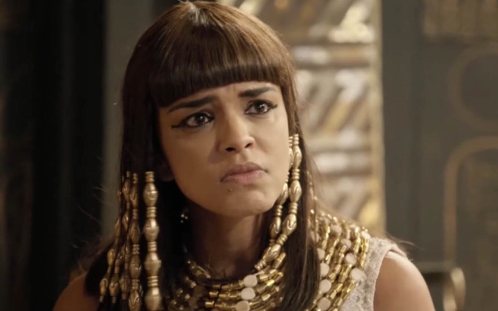 A atriz Kizi Vaz com uma peruca de fios negros, de franja, adornada com contas está caracterizada como Kamesha com expressão de fúria em cena de Gênesis