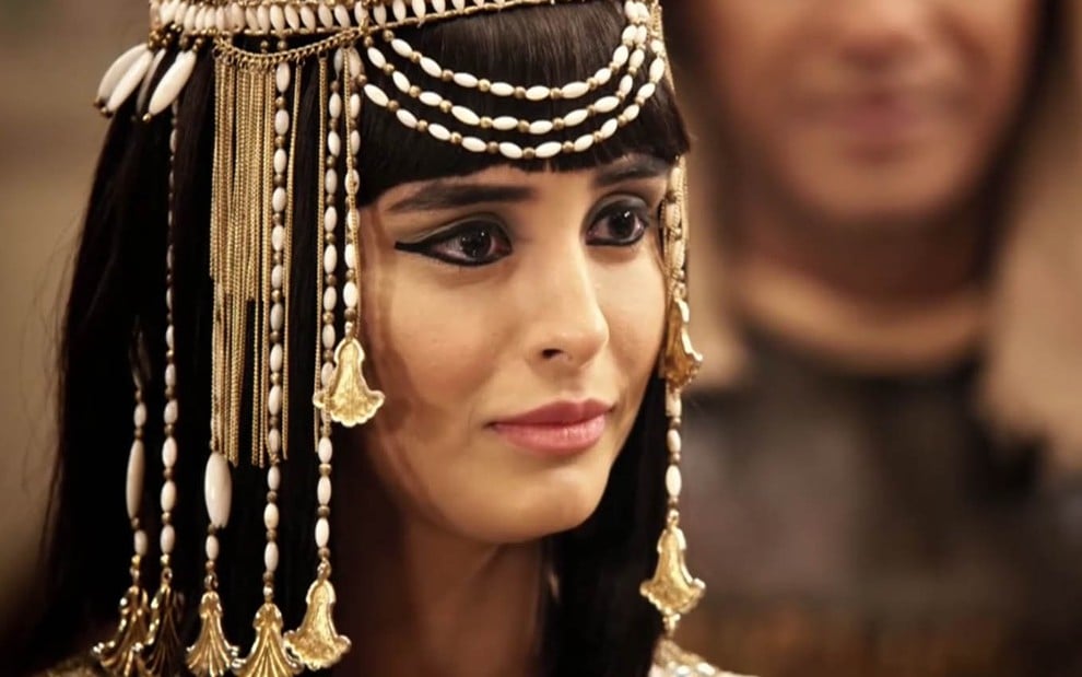 A atriz Letícia Almeida em um close no seu rosto, com uma peruca de fios pretos adornada com ouro, caracterizada como Asenate em cena de Gênesis