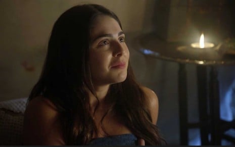 A atriz Marcela Barrozo está nua, com um lençol cobrindo o corpo, enquanto está sentada em uma cama com os cabelos pretos, lisos e soltos como a Uriala em cena de Gênesis