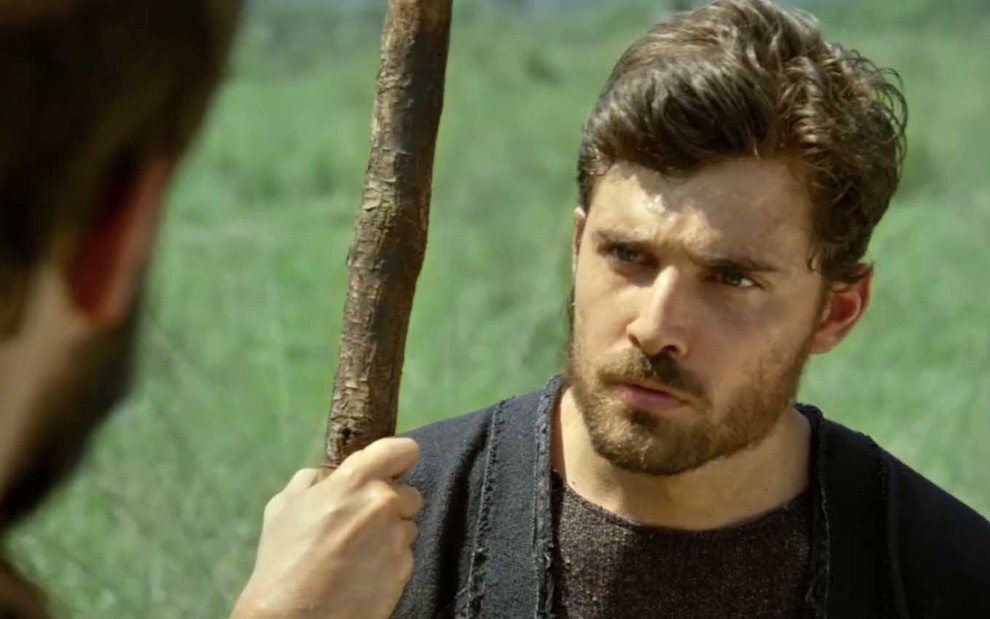 O ator Miguel Coelho está em um pasto muito verde e segura um cajado de madeira escura com a mão direita com expressão de raiva como Jacó em cena de Gênesis