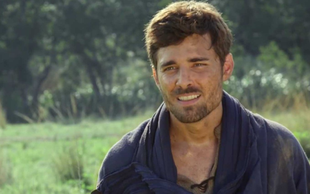 O ator Miguel Coelho caracterizado como Jacó está com o rosto sujo de terra em um pasto verde, com uma pequena floresta ao fundo, em uma cena ensolarada de Gênesis
