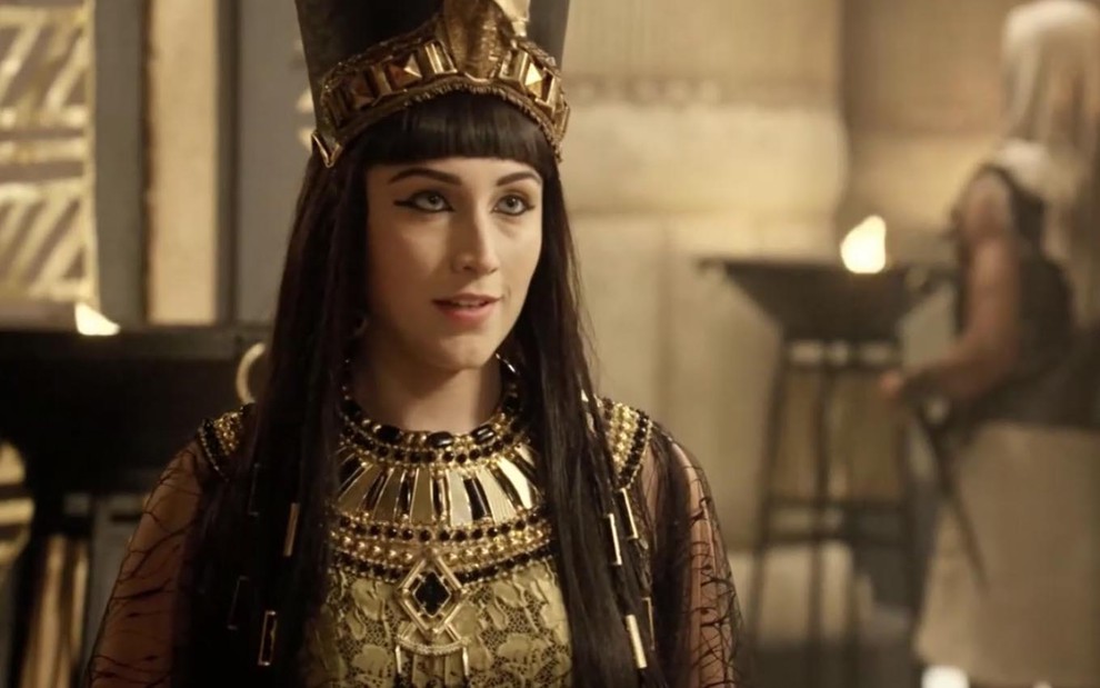 A atriz Samia Abreu está com uma roupa ricamente adornada com detalhes dourados e um chapéu ao estilo Nefertari como a Merianat em cena de Gênesis