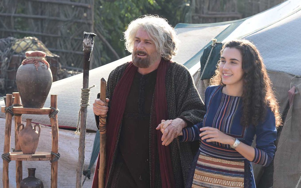 Diante de uma tenda, o ator Petrônio Gontijo como Israel é conduzido por Giovanna Coimbra, a Diná, enquanto se apoia em um cajado em cena de Gênesis