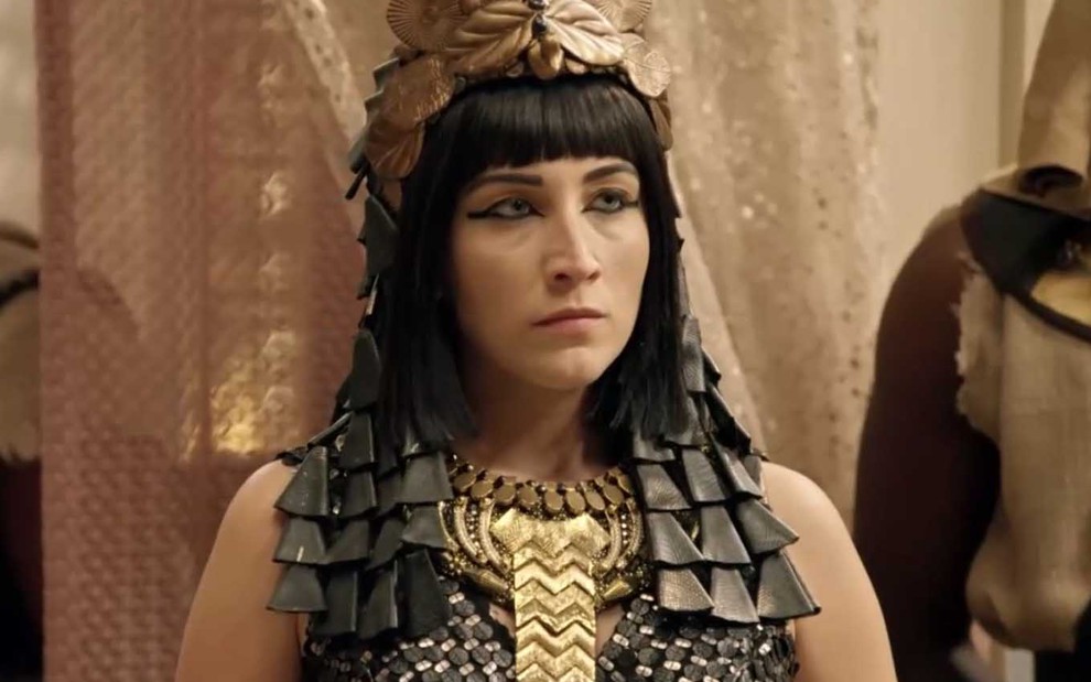 A atriz Smaia Abreu está com uma peruca preta, de franja, adornada com flores de cobre e placas de metal como a rainha Merianat em cena de Gênesis