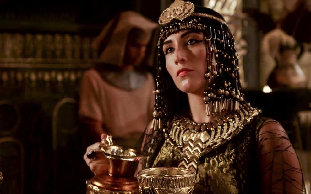 A atriz Samia Abreu caracterizada como Merianat, com o figurino adornado de ouro, segurando uma taça e uma jarra de vinho em cena de Gênesis
