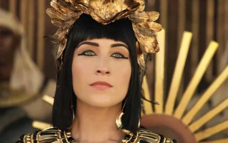 A atri Samia Abreu está diante de um trono adornado com ouro, com uma peruca de fios pretos, de franja, com uma peça em penas de pardal e ouro na cabeça como a Merianat em cena de Gênesis