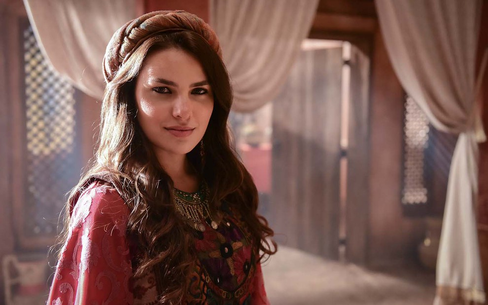 A atriz Thaís Melchior está com um vestido vermelho, longo, e um turbante no topo da cabeça que permite aos seus cabelos ficarem soltos como a Raquel em cena de Gênesis
