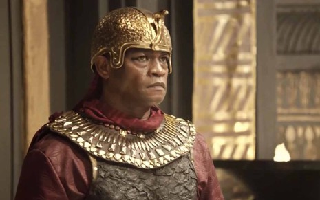 O ator Val Perré está com um elmo de guerreiro dourado, uma capa vermelha e um peitoral com detalhes dourados como o general Potifar em cena de Gênesis