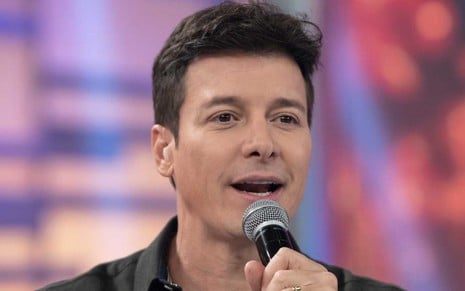 O apresentador Rodrigo Faro em um close no seu rosto, ele segura um microfone com a mão direita