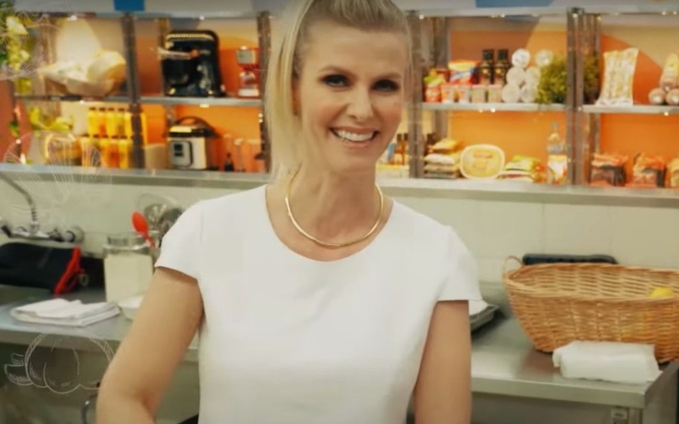 Apresentadora loira, Thays Beleze veste camiseta branca e está em uma cozinha com equipamentos ao fundo