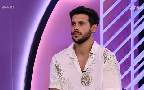 Rodrigo Mussi de camisa de botão branca em cenário roxo e lilás dos estúdios da Rede BBB