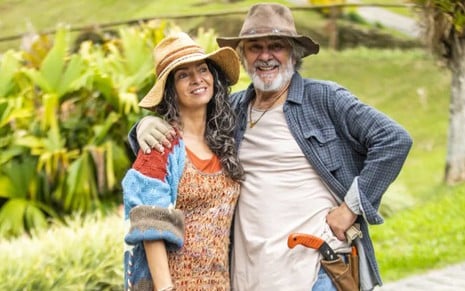 Os atores Claudia Ohana e Zécarlos Machado abraçados, lado a lado, sorrindo, em paisagem de campo, em cena de Vai na Fé