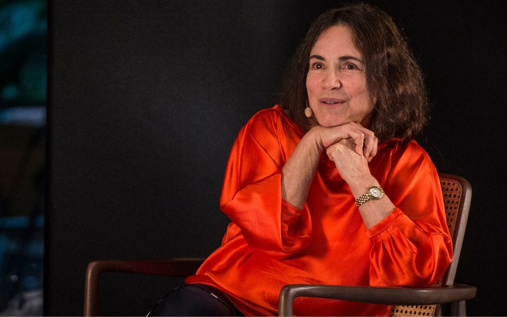 Em cenário de teatro preto, Regina Duarte faz cara reflexiva sentada em cadeira marrom