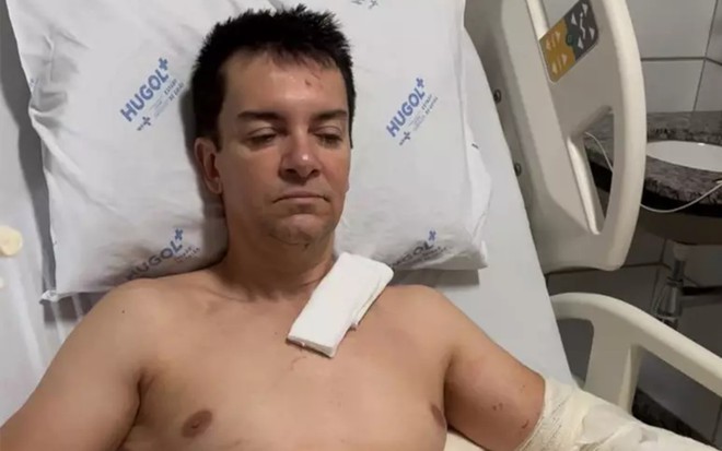 Regis Danese está de olhos entreabertos, sem camisa, deitado na cama de um hospital