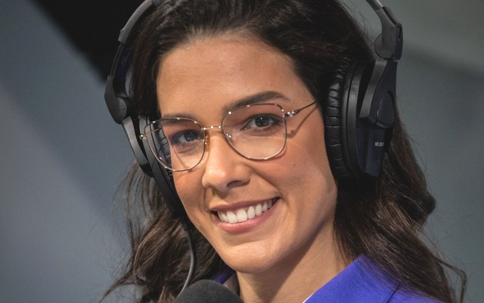 Renata Silveira usa o uniforme azul da Globo nos bastidores de transmissão esportiva; ela está sorridente