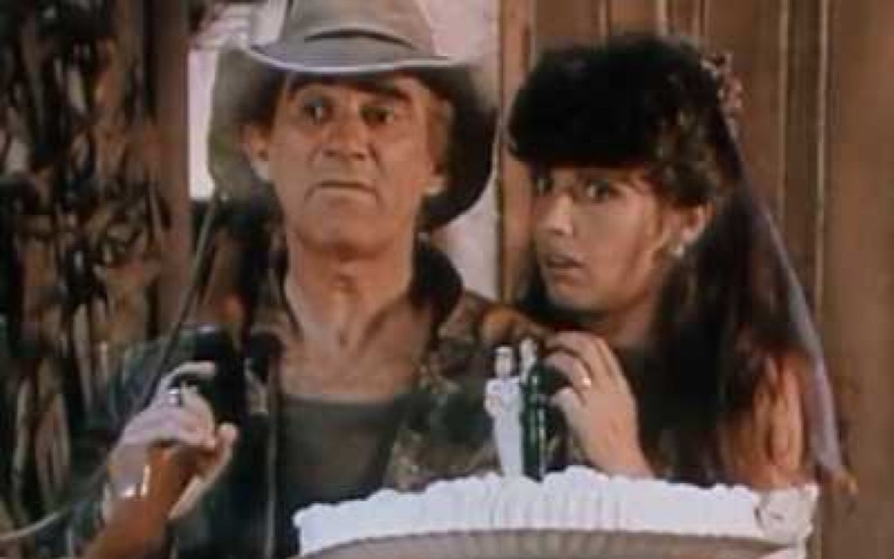 Renato Aragão com um bolo de casamento na mão, segurando uma arma, vestindo um chapéu; ao seu lado está Nádia Lippi apoiada em seu ombro, com uma cara de assustada