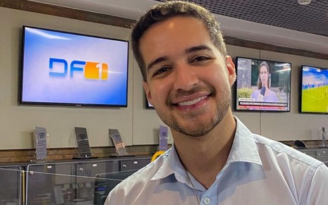 Repórter Gabriel Luiz veste camisa social e sorri com TVs ao fundo na Redação da Globo em Brasília