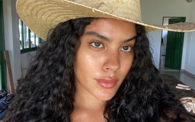 A atriz Bella Campos está com o cabelo solto e usa um chapéu de palha