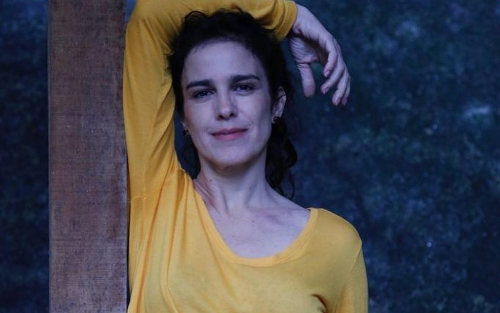 A atriz Karina Barum, com uma blusa amarela, com o braço apoiado na cabeça