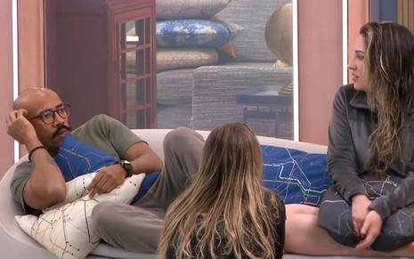Ricardo Camargo deitado no sofá da área externa ao lado de Amanda Meirelles e Bruna Griphao, que está sentada no chão