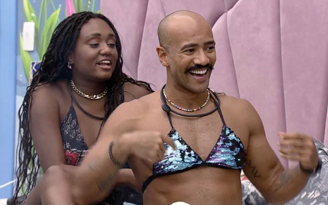 Ricardo Camargo usa um biquíni de paetês verde; Sarah Aline está atrás dele, rindo, no BBB 23
