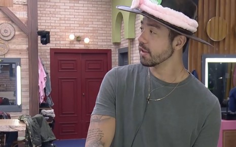 Na sede de A Fazenda, Rico veste camiseta cinza e usa chapéu de fazendeiro com faixa de plumas rosa