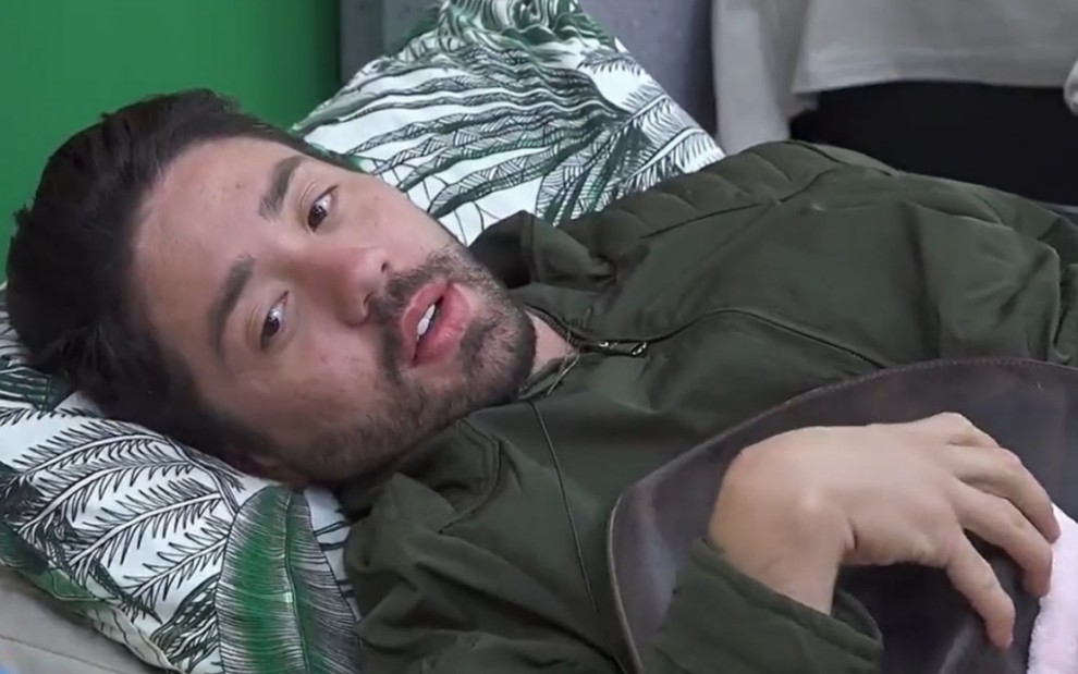 O influenciador Rico Melquiades com uma jaqueta verde, deitado na cama, com olhar distante em A Fazenda 13