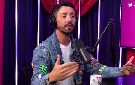 Rico Melquiades no podcast Podcats, ele está sentado próximo a um microfone e veste uma camiseta branca e uma jaqueta jeans