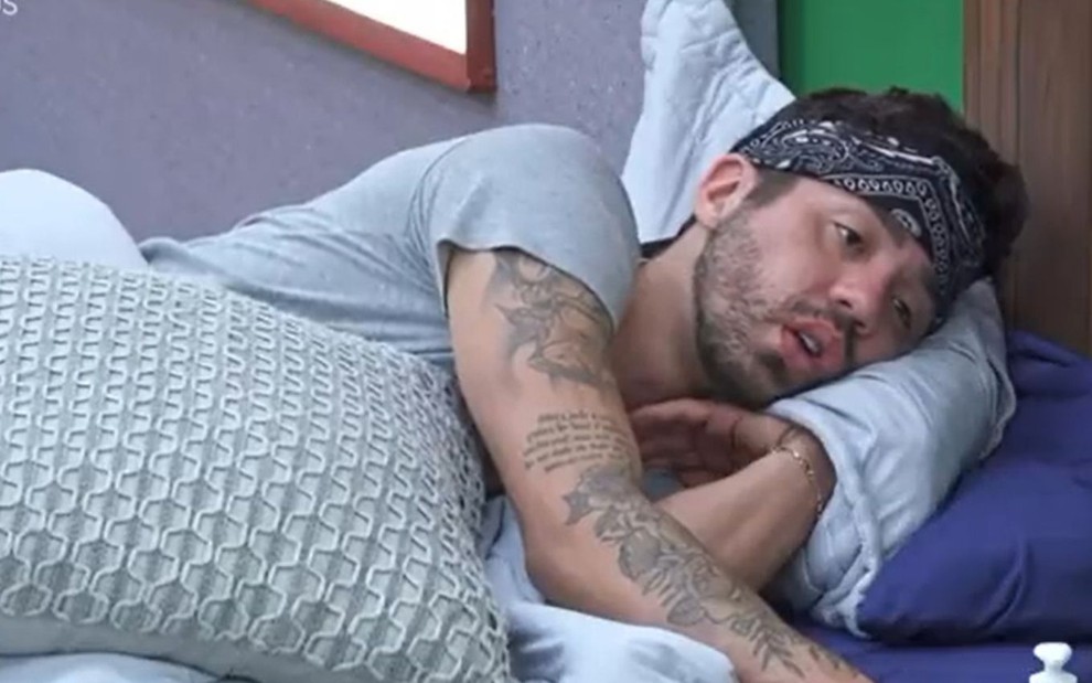 Rico Melquiades de bandana preta na testa, deitado na cama de camiseta cinza e expõe tatuagens no braço direito