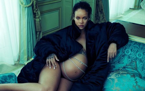Rihanna sentada no sofá, vestindo uma jaqueta e com a barriga à mostra