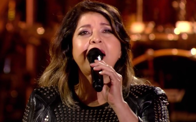 Roberta olha para cima, veste um vestido escuro e segura um microfone