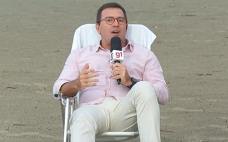 Rodrigo Bocardi sentado em uma cadeira de praia na areia e segurando um microfone