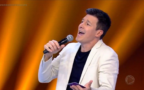 Rodrigo Faro usa um blazer branco com blusa preta e sorri para o Paredão dos Famosos, quadro de seu programa nos domingos da Record