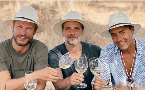 Rodrigo Hilbert, Bruno Gagliasso e Ricardo Pereira estão de chapéu e brindando com taças de vinho