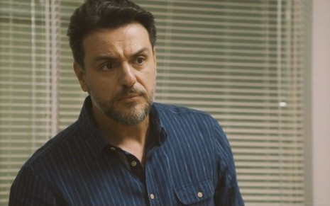 Em cena de Travessia, Rodrigo Lombardi está usando uma blusa azul com listras finas brancas e está com a expressão de descontentamento