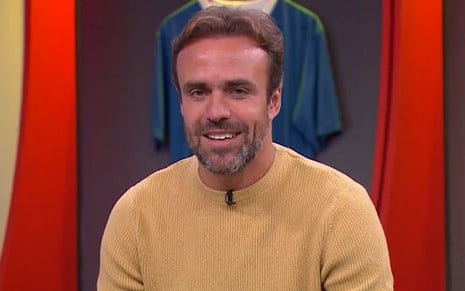 Roger Flores sorri discretamente e usa uma blusa mostarda