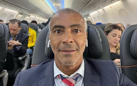Romário está sentado numa poltrona de avião e encara a câmera; ele usa um terno grafite e fones de ouvido brancos.