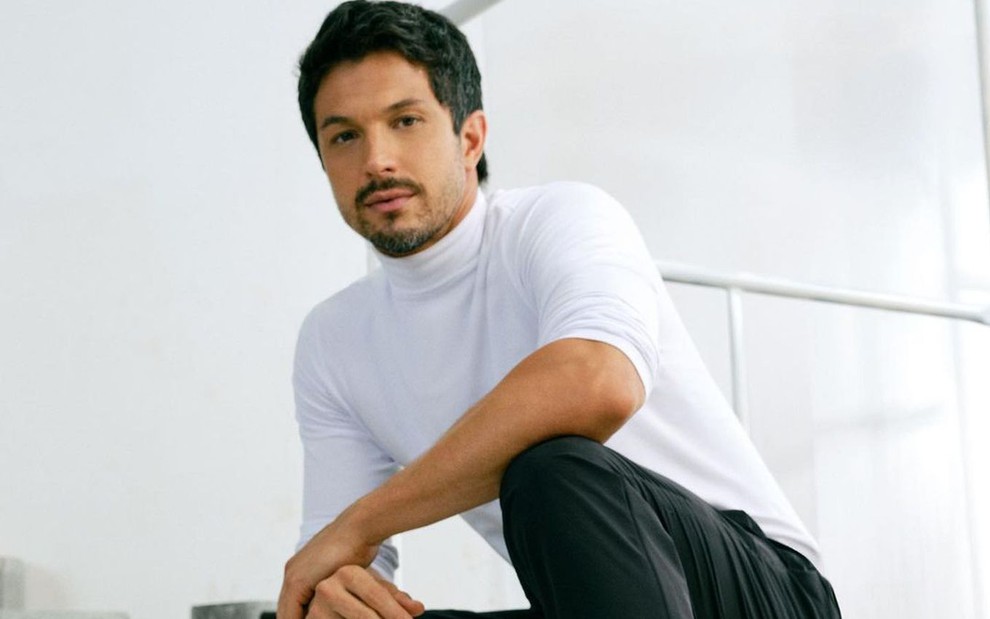 O ator Romulo Estrela está posando para a câmera segurando um dos braços e agachado no chão