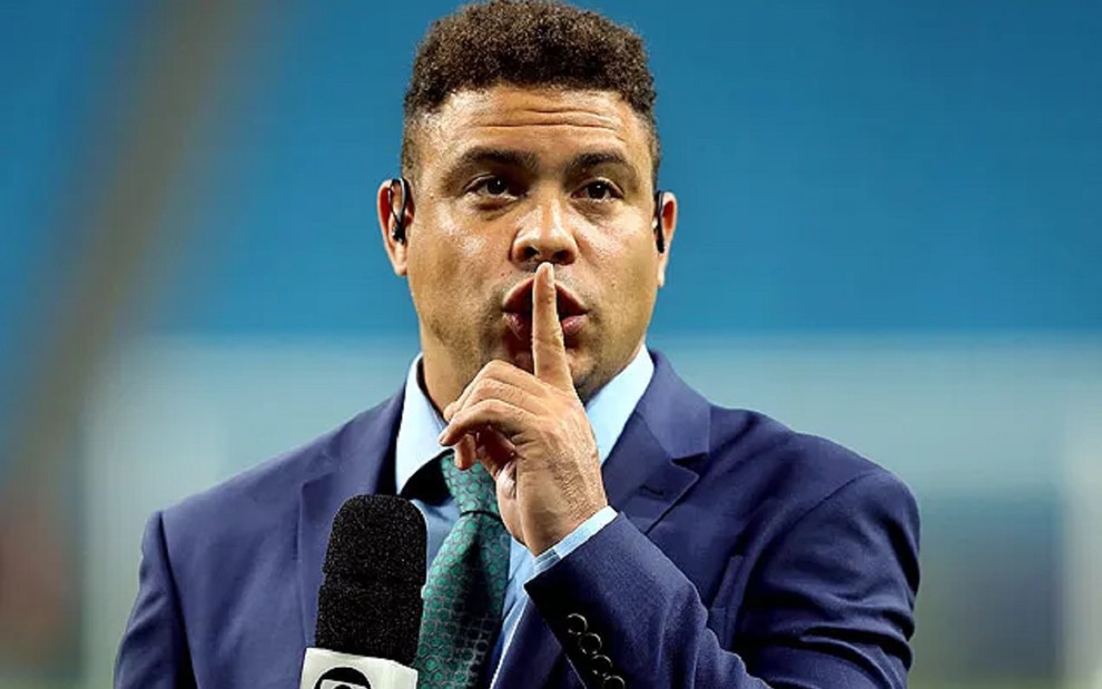 Ronaldo Fenômeno com um terno azul, gravata verde e um sinal de silêncio durante cobertura de um jogo na Seleção Brasileira