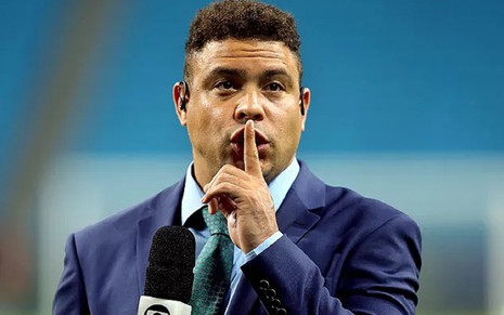 Ronaldo Fenômeno com um terno azul, gravata verde e um sinal de silêncio durante cobertura de um jogo na Seleção Brasileira