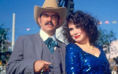 Sinhozinho Malta (Lima Duarte) e viúva Porcina (Regina Duarte) lado a lado em cena externa Roque Santeiro; ele de terno, gravata e chapéu, ela de vestido azul brilhante